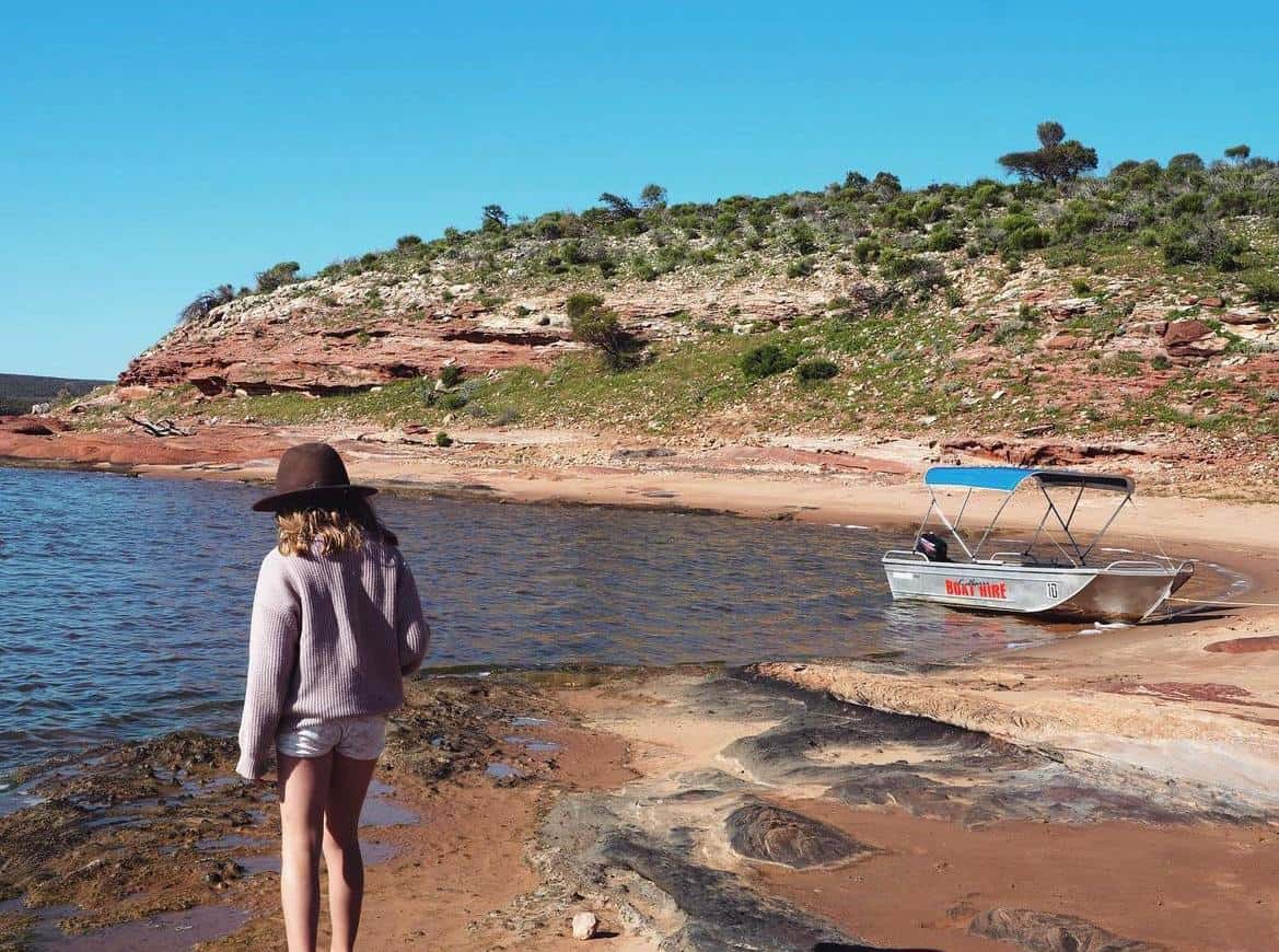 A girl in a casual outfit and a wide-brimmed hat stands on a sandy shore, gazing out at a moored boat with 'Boat Hire' text, set against the stunning backdrop of the layered cliffs and clear waters near Kalbarri, Western Australia.