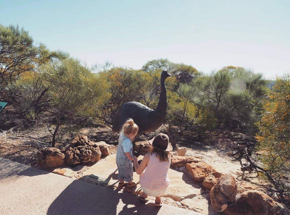 A young child and an adult, seen from behind, admiring a large emu statue amidst the lush bushland, a moment of exploration and learning about native wildlife on a sunny day in Kalbarri, Western Australia.