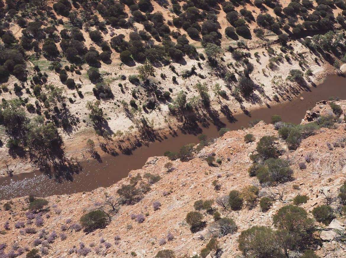 Aerial view of a meandering river cutting through a rugged landscape with sparse vegetation, captured on the journey from Perth to Kalbarri in Western Australia, highlighting the remote beauty of the region.