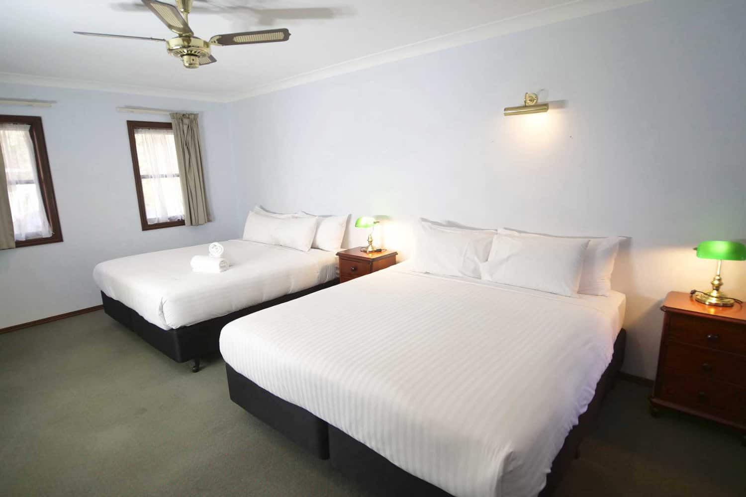 A well-lit, clean hotel room with two queen-sized beds, white bedding, side tables with lamps, and a ceiling fan, offering a cozy and inviting atmosphere.
