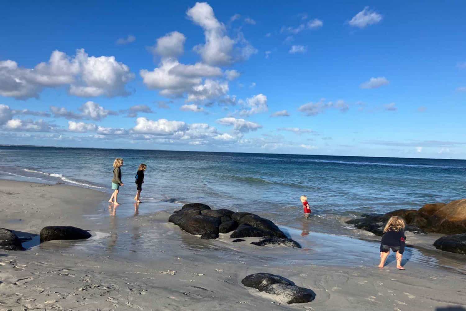 Children wade and play near the water's edge on an Augusta beach, Dead Finish, with natural black rocks embedded in the sand, under a sky dotted with cotton-like clouds, offering a snapshot of family moments by the sea
