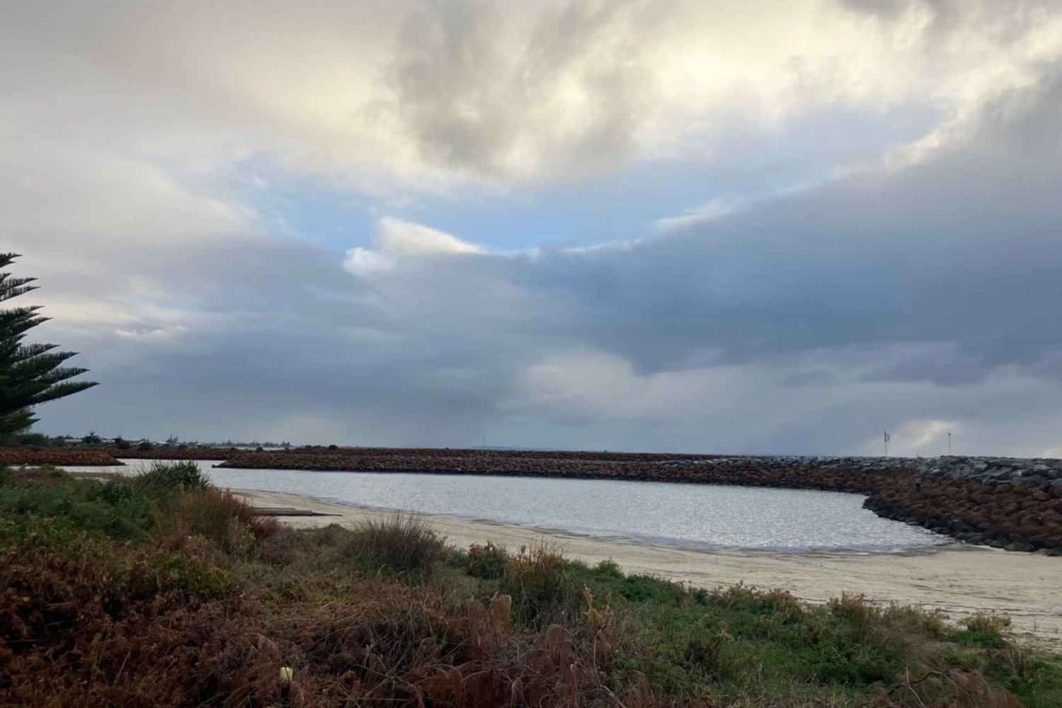 Moody skies over the calm inlet at Busselton beaches, with a protective breakwater and coastal vegetation.