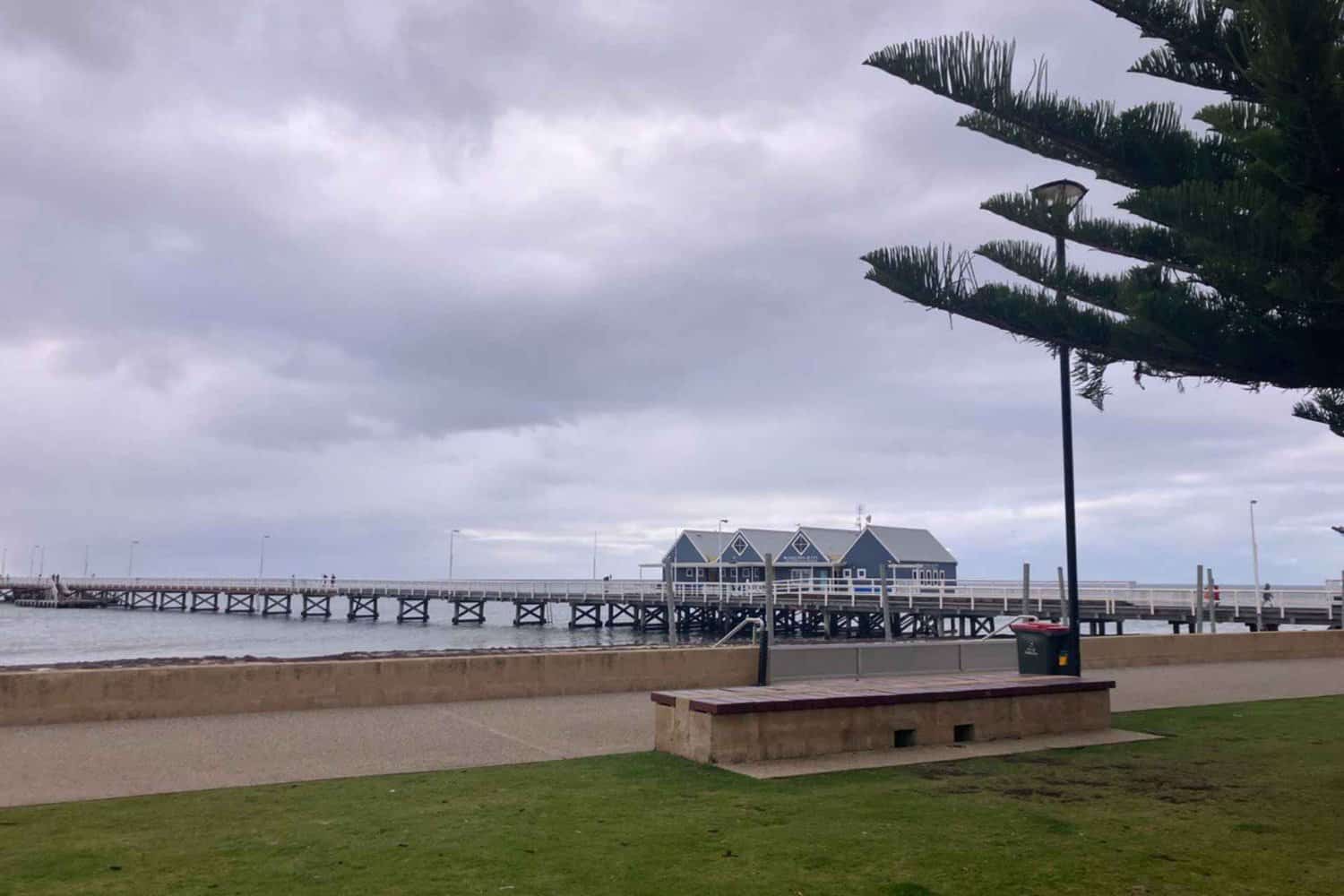 Gloomy weather over the iconic Busselton Jetty extending into the sea, with a lush park area in the foreground.