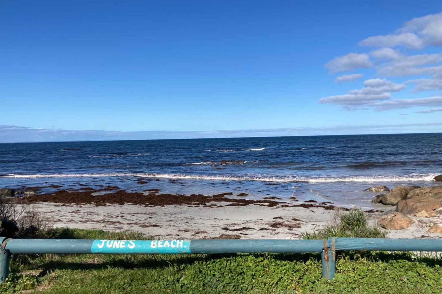 The rustic charm of Jay's Beach in Augusta is marked by a simple green barrier with the beach's name, leading the eye to the rocky shoreline and gentle waves, set against the vast expanse of the ocean and a clear blue sky with scattered clouds.