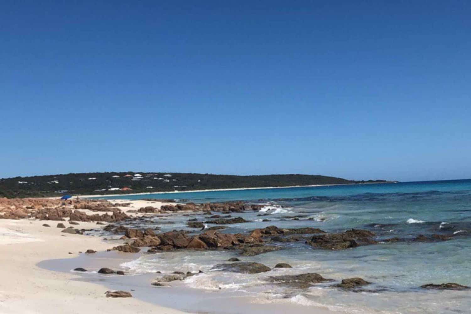 Serene Dunsborough dog beach with clear blue skies, gentle waves lapping over scattered rocks, and a pristine stretch of sand, inviting dog owners for a picturesque walk with their pets.