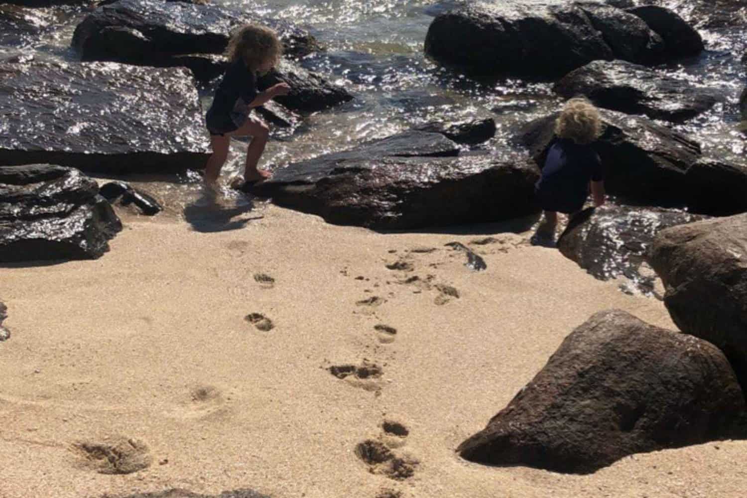 Children explore the natural rock pools on a sunlit Margaret River beach, leaving a trail of footprints in the sand, capturing a moment of discovery and play on the rugged coastline.