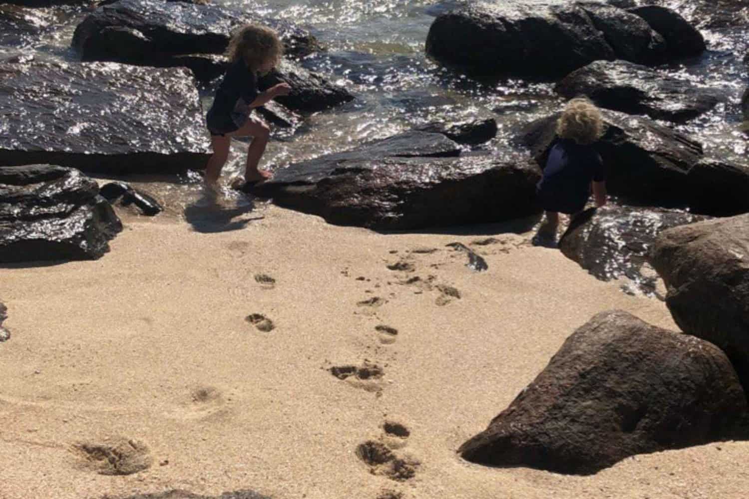 Two children exploring a sandy beach with footprints trailing behind them, playing between the rocks and splashing water in a moment of joy and discovery.