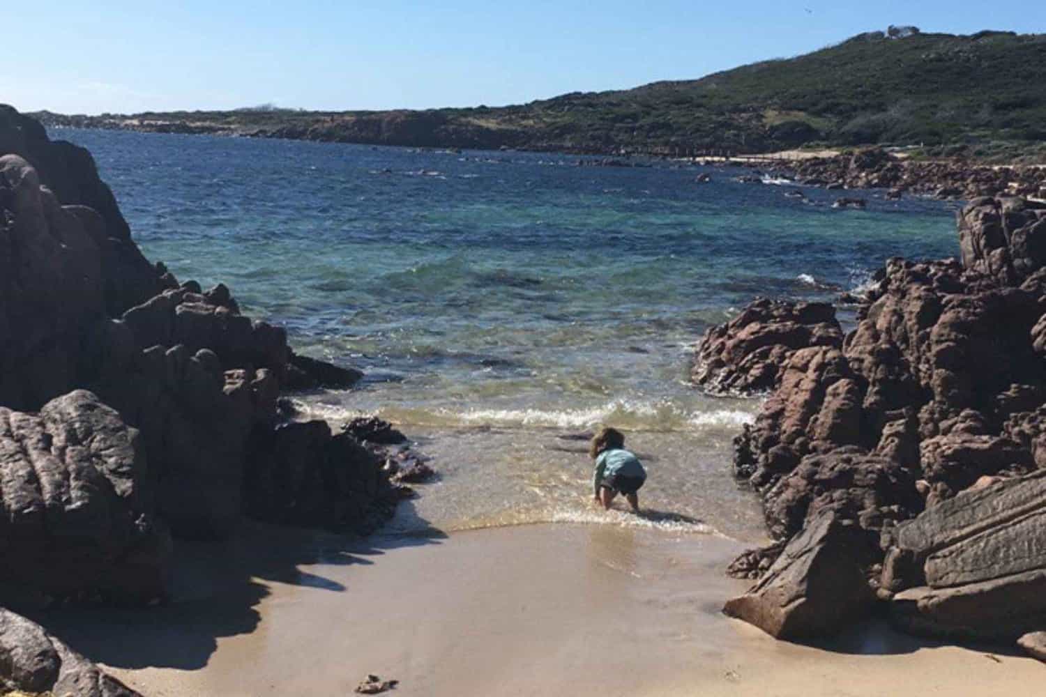 A child crouches on the sandy shore of a Gracetown beach, exploring the water's edge where the sea meets a cluster of dark, weathered rocks, with the peaceful ocean extending to the horizon under a sky dotted with fluffy clouds.