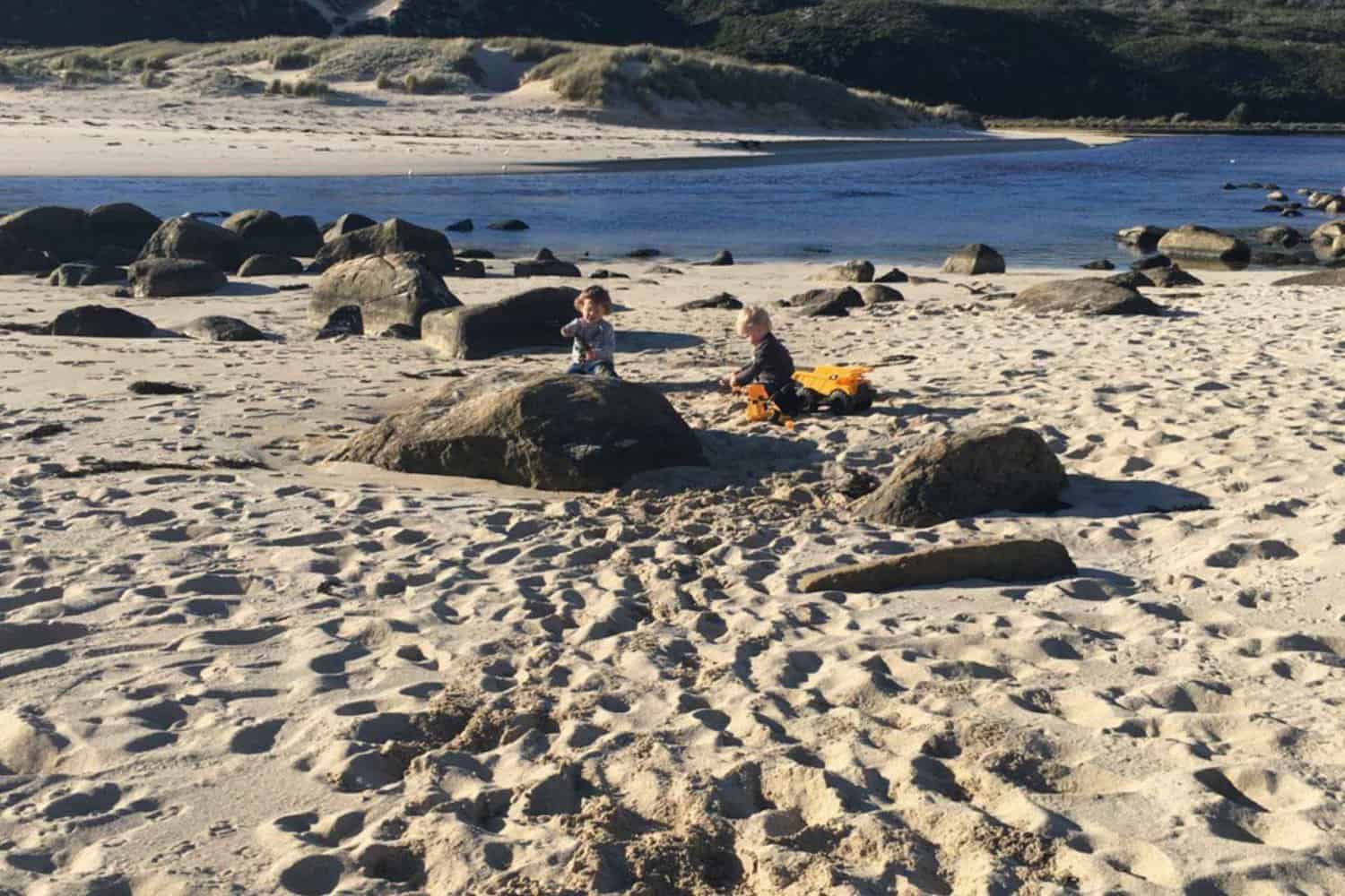 Children play in the sand surrounded by natural rock formations on a Margaret River beach, with the calm sea and sand dunes under the soft glow of the late afternoon sun, encapsulating the serene and family-friendly atmosphere.