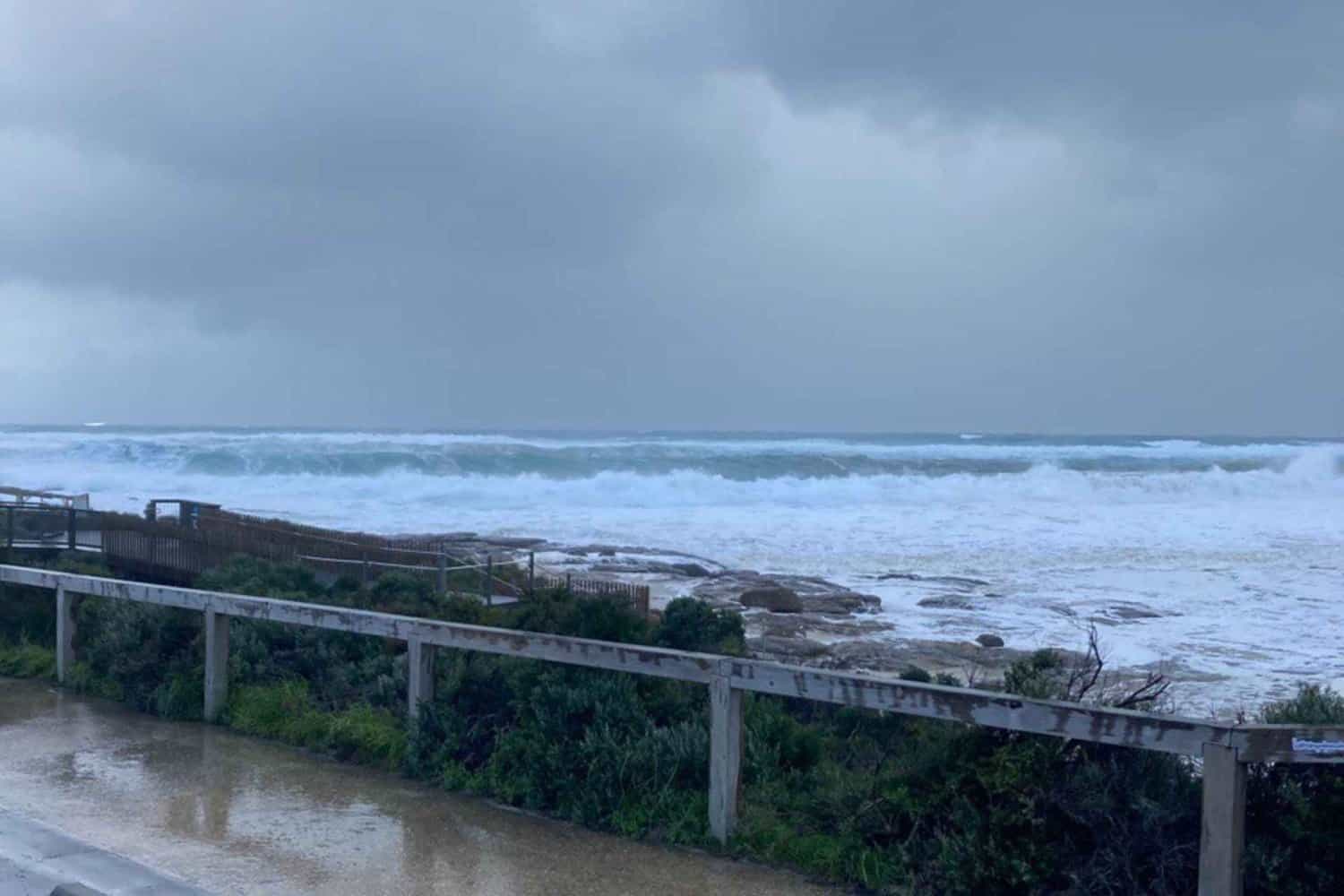 A stormy sky casts a dramatic backdrop over the rough, foamy waves of a Margaret River beach, as seen from a coastal walkway lined with a wooden railing, reflecting the beach's wild beauty during tumultuous weather