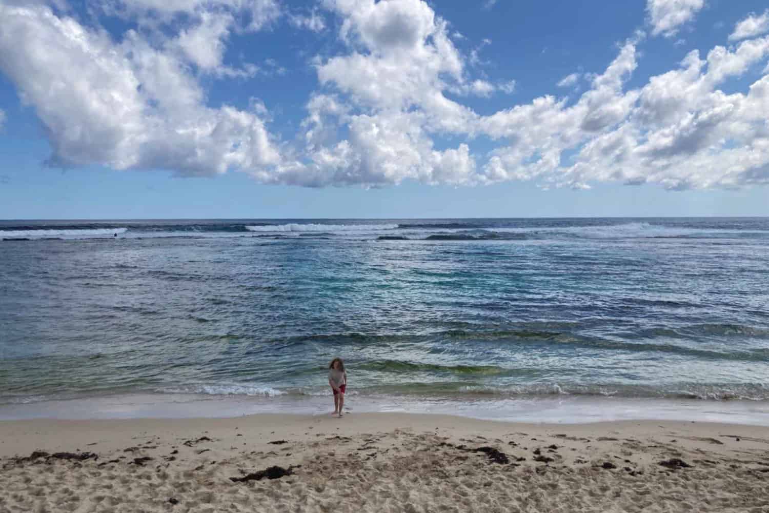 An afternoon at Yallingup Dog Beach with a small child standing at the water's edge, facing the expansive view of rolling waves under a sky filled with dynamic cloud formations.