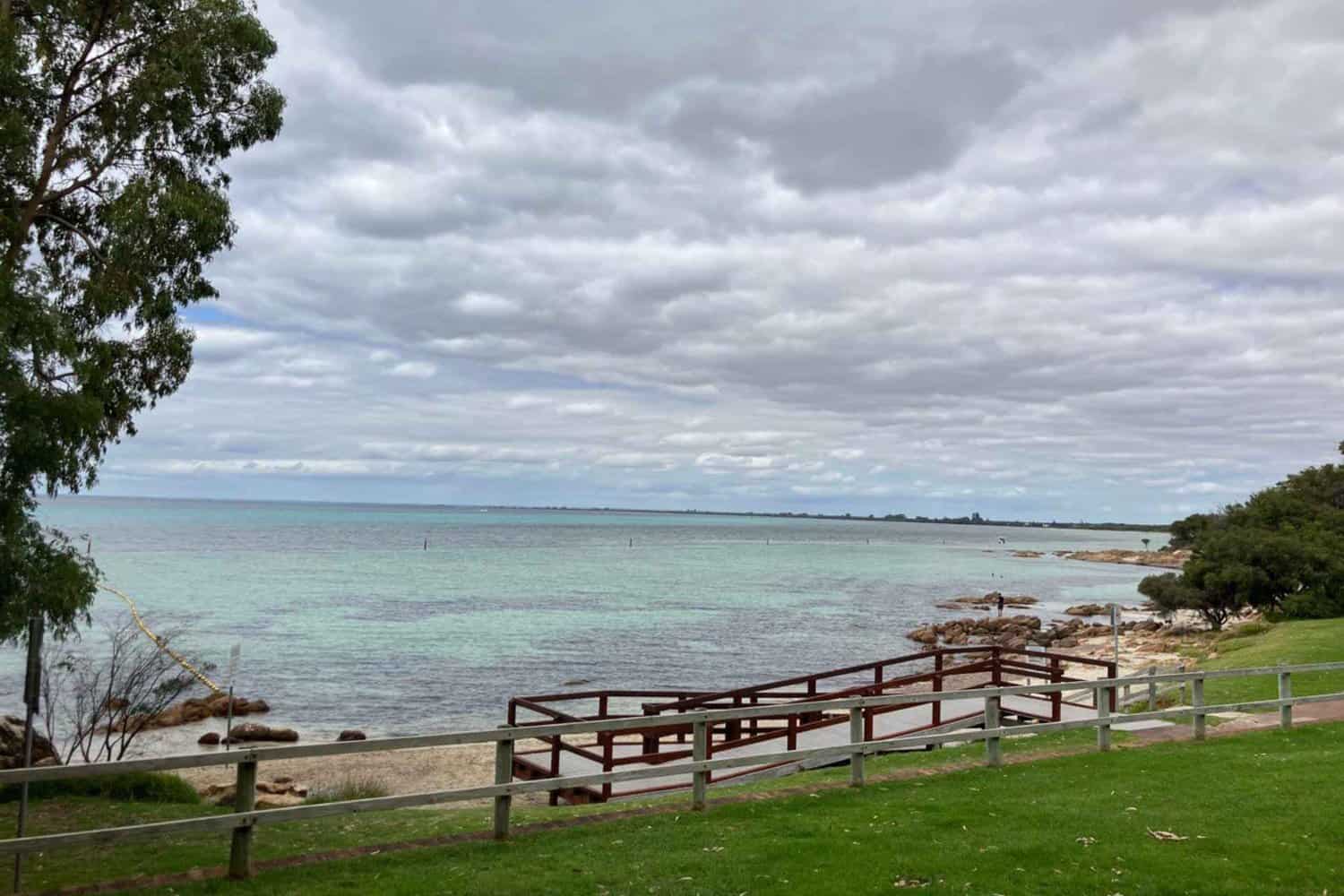 Overcast skies above the turquoise waters of Dunsborough beaches, with a lush green park and wooden fence in the foreground.