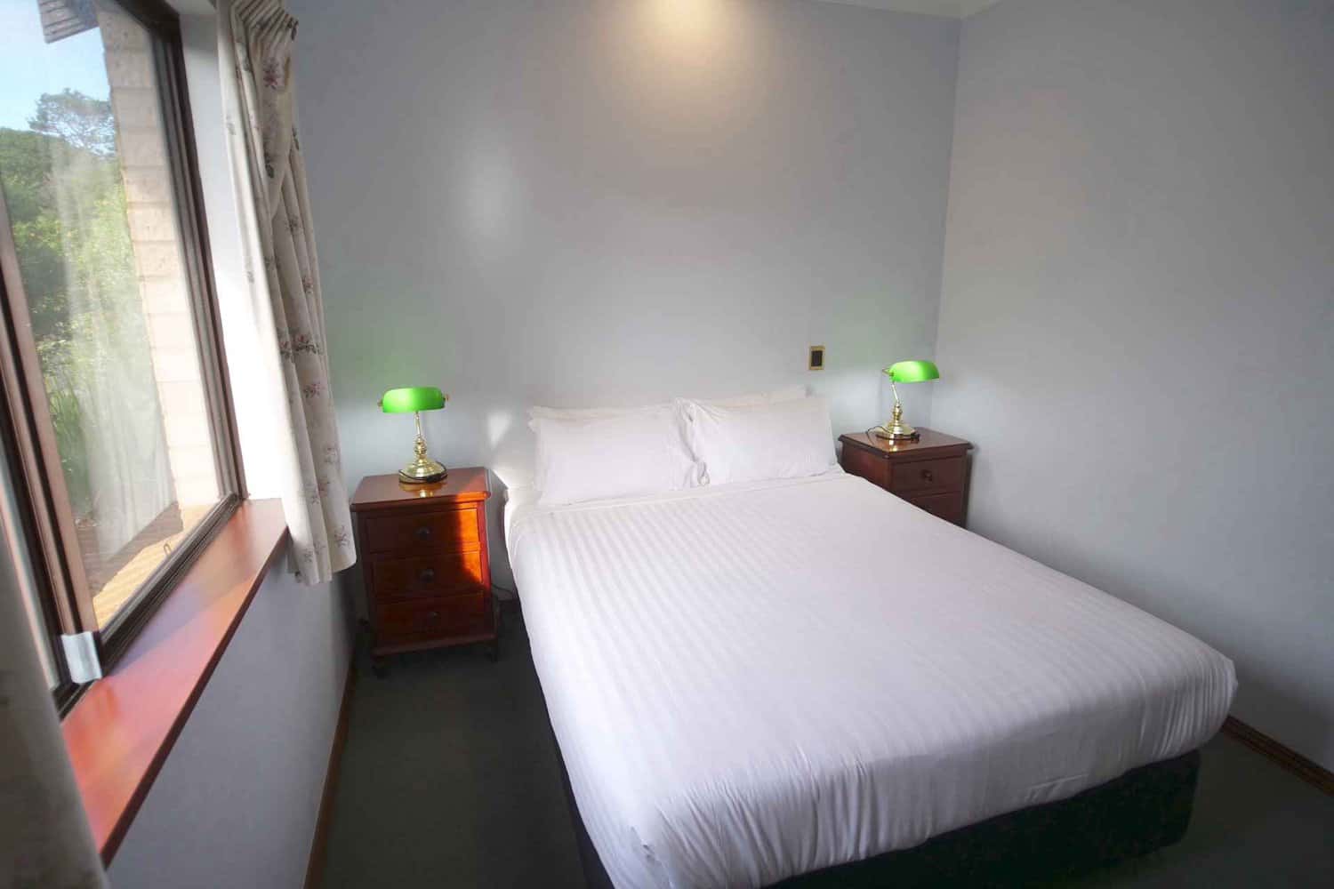 Minimalist hotel room with a comfortable single bed, clean white bedding, and matching wooden nightstands with green shaded lamps, next to a window with light curtains and a view of greenery outside.