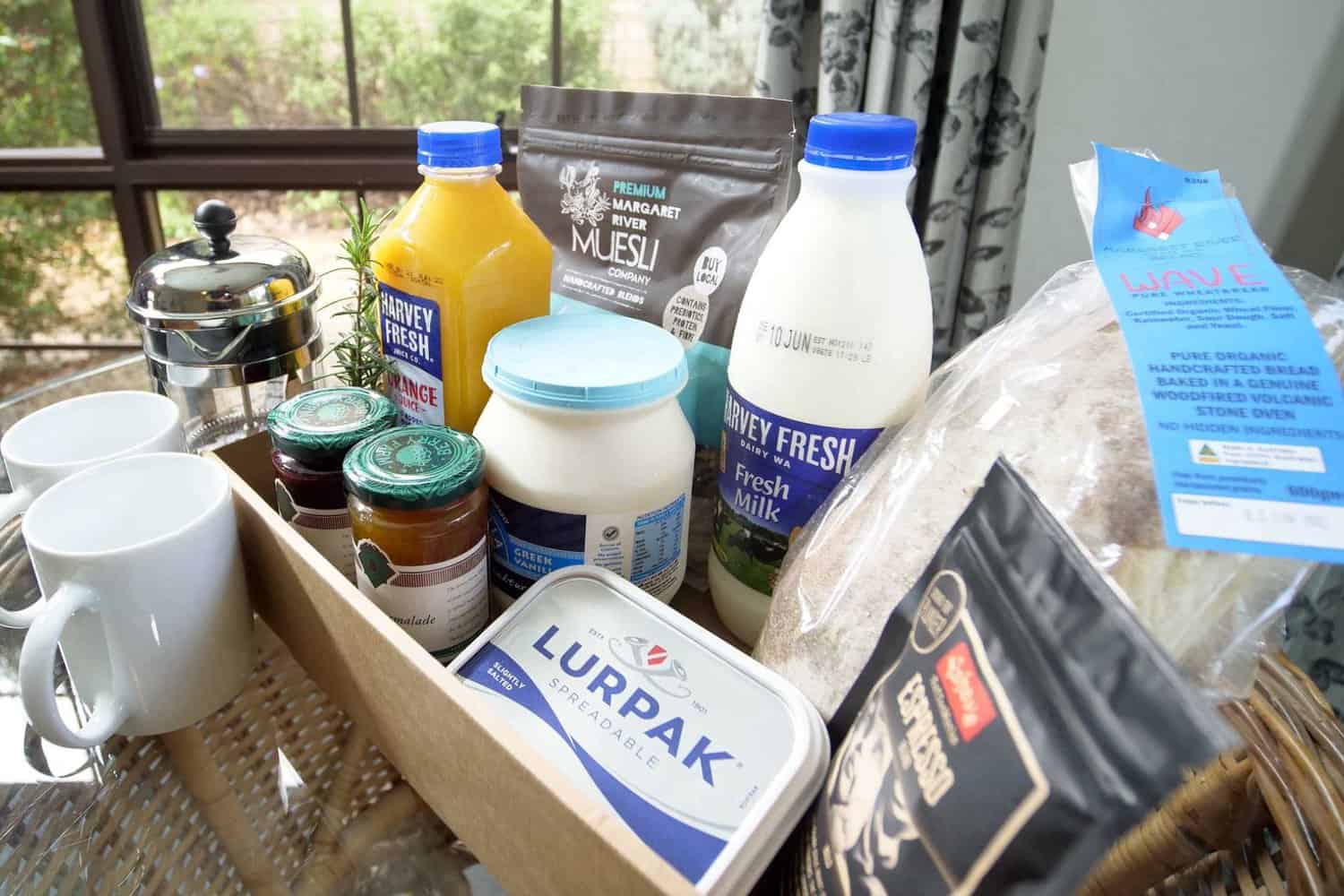 A tray with a variety of breakfast essentials including Harvey Fresh orange juice, natural yogurt, muesli, fresh milk, artisan bread, Lurpak butter, gourmet jams, and a bag of espresso coffee, ready for a nourishing start to the day.