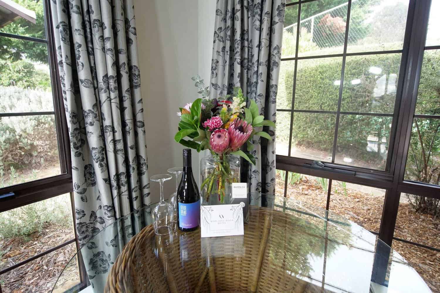 An inviting hotel room setting with a bottle of red wine and two glasses on a round wicker table, featuring a lush bouquet of native flowers and a Margaret River Motel leaflet, with garden views through the window.
