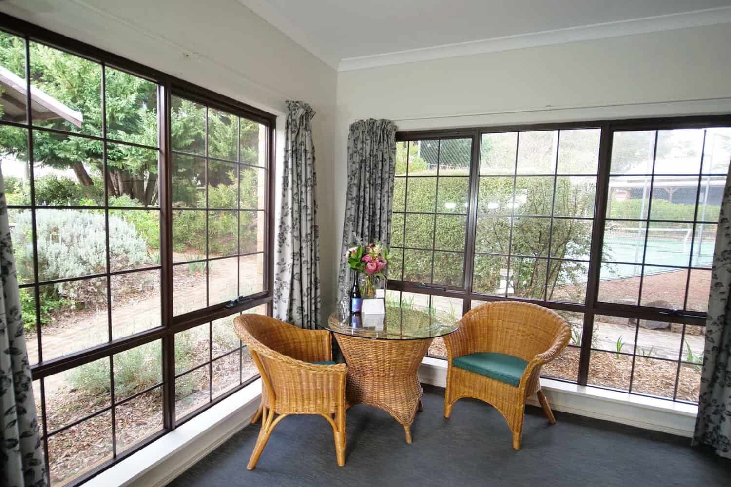 A cozy hotel room corner with a glass table and wicker chairs by a large window, offering a view of the peaceful garden outside, perfect for a relaxing morning or afternoon.