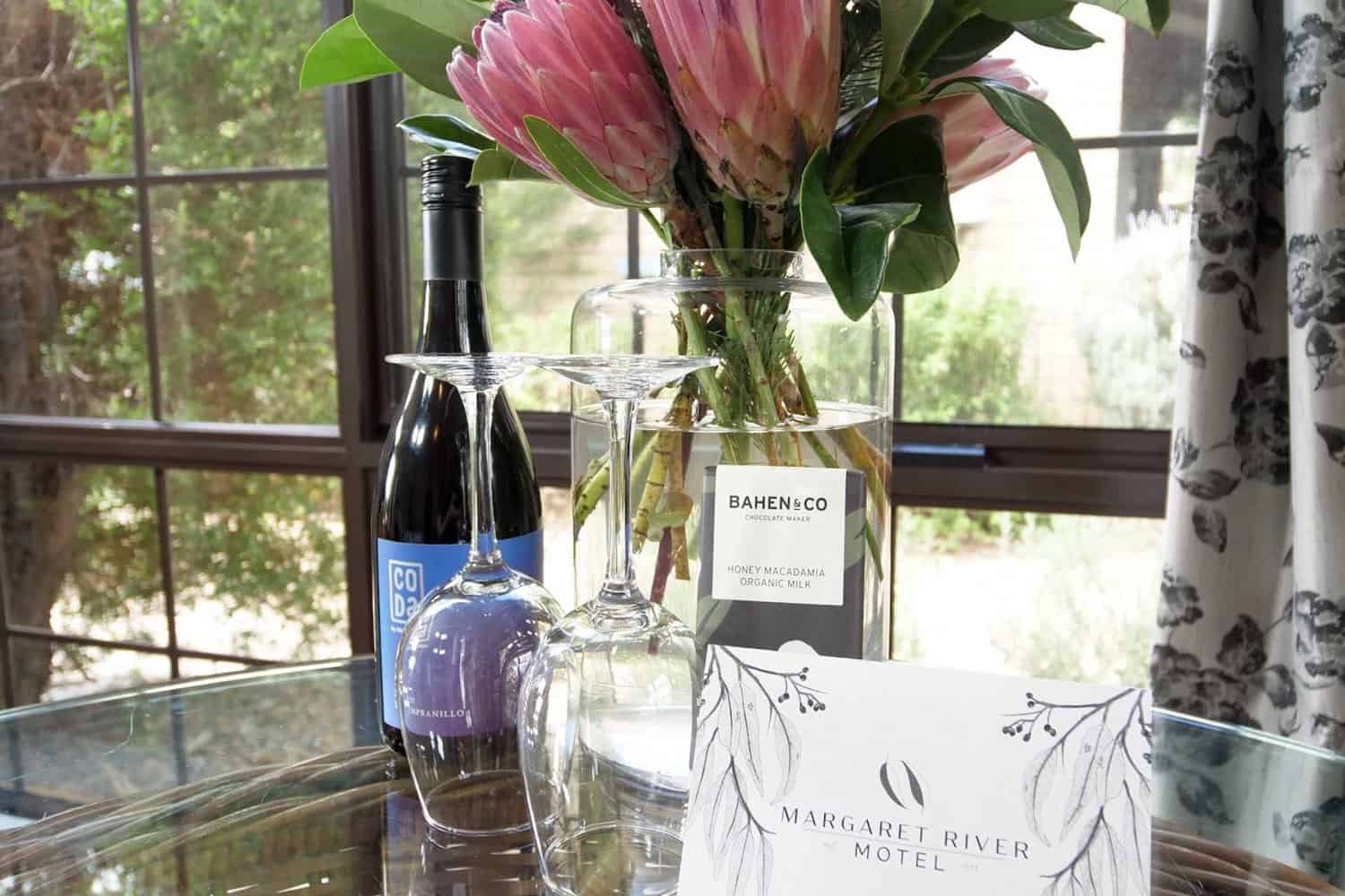 A welcoming hotel room scene with a bottle of red wine and two glasses set on a glass table, accompanied by a vibrant pink protea bouquet and a bar of Bahen & Co. honey macadamia chocolate, with a 'Margaret River Motel' pamphlet suggesting local flair.
