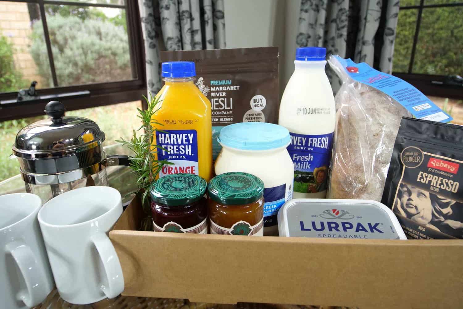 A selection of breakfast items including fresh orange juice, milk, muesli, yogurt, jams, butter, and espresso coffee, all arranged neatly in a box, ready for a delightful morning meal in a room with garden view curtains.