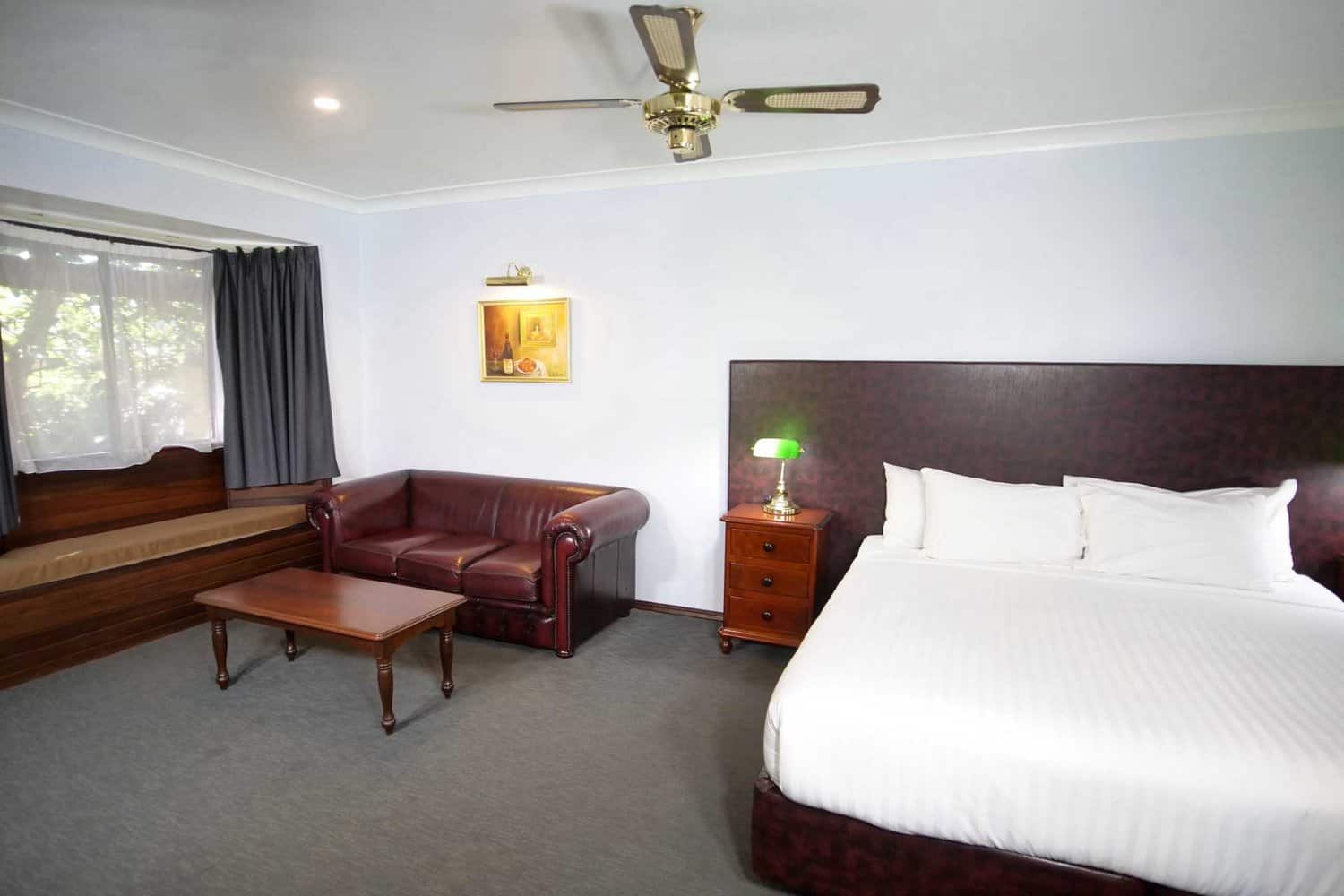 Classic hotel room with a large comfortable bed with white linens, a leather sofa, and a wooden coffee table, complete with a ceiling fan and a soft-lit lamp, providing a cozy atmosphere for guests.