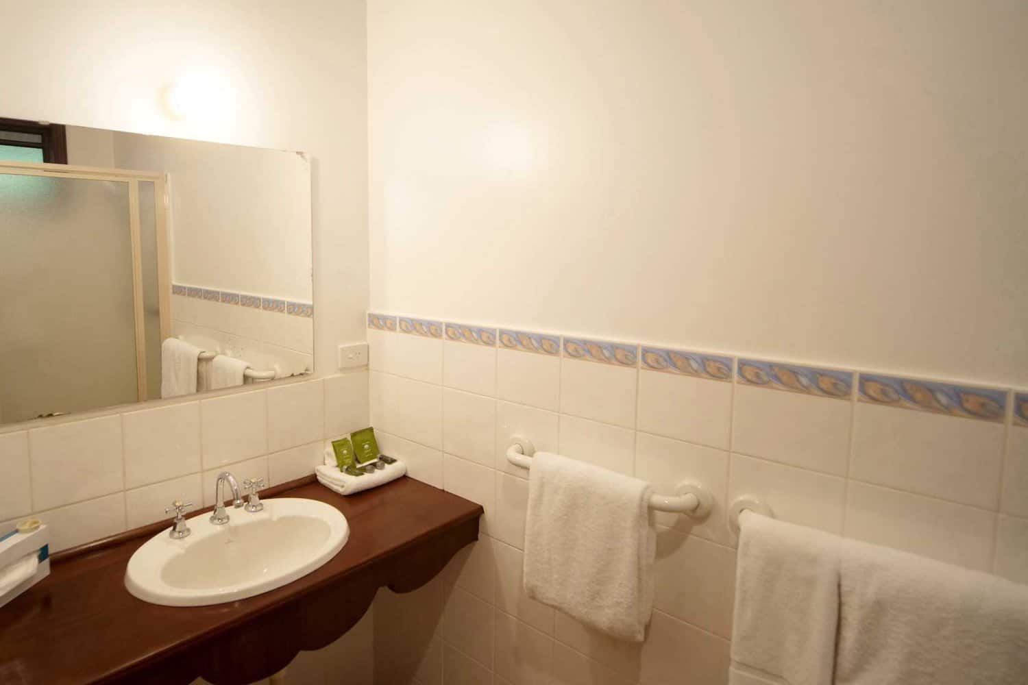 Clean and simple hotel bathroom with a large mirror, white tiled walls featuring a blue tile border, a wooden vanity with a white basin, and fluffy towels ready for use.