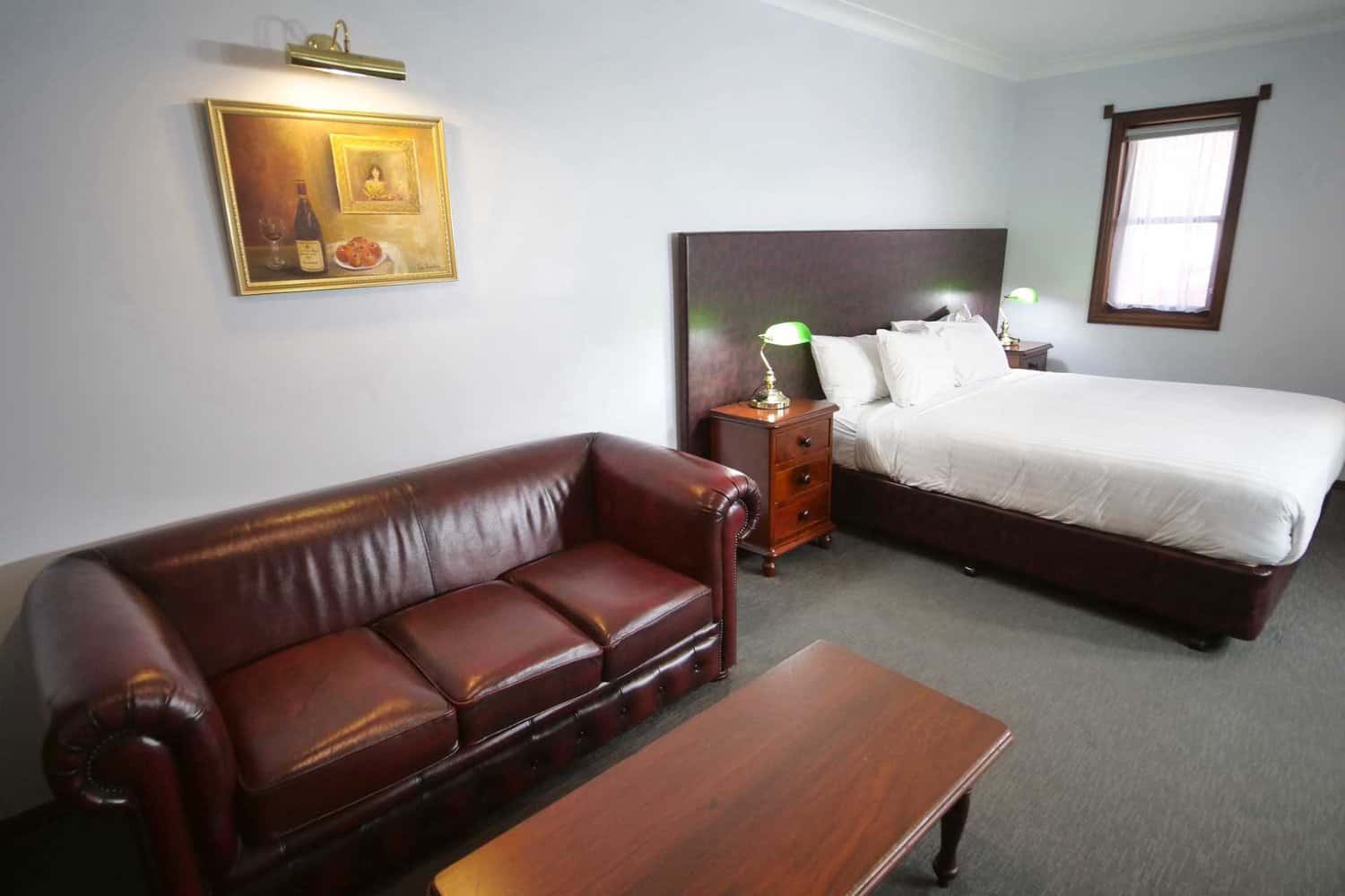 A spacious hotel room elegantly arranged with a large bed topped with white linens, a classic leather sofa, and a polished coffee table, under the warm light from green-shaded lamps, next to a window with natural light.