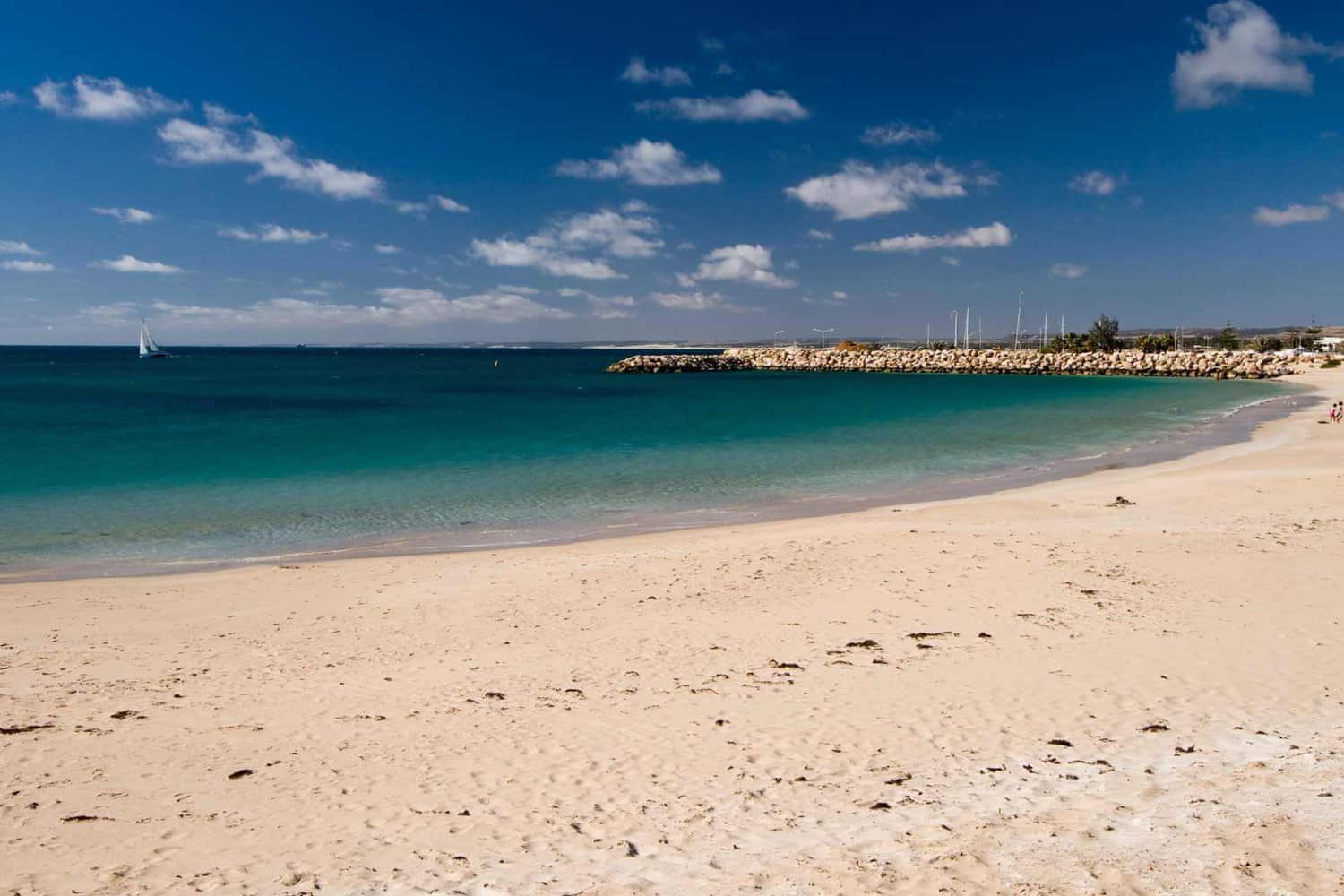 A tranquil beach scene with clear turquoise waters and a sailboat in the distance, complemented by a sunny blue sky dotted with fluffy clouds. The sandy shoreline and protective rock wall create a picturesque setting near Geraldton.