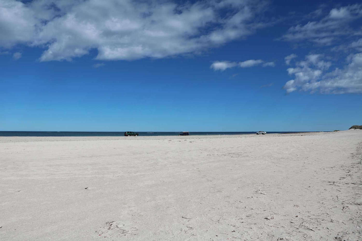A vast, white sandy beach extends to the calm blue ocean under a sky scattered with clouds, with a few 4WD vehicles parked in the distance. This spacious beach setting is perfect for adventurers seeking free camping spots along the Geraldton coast.