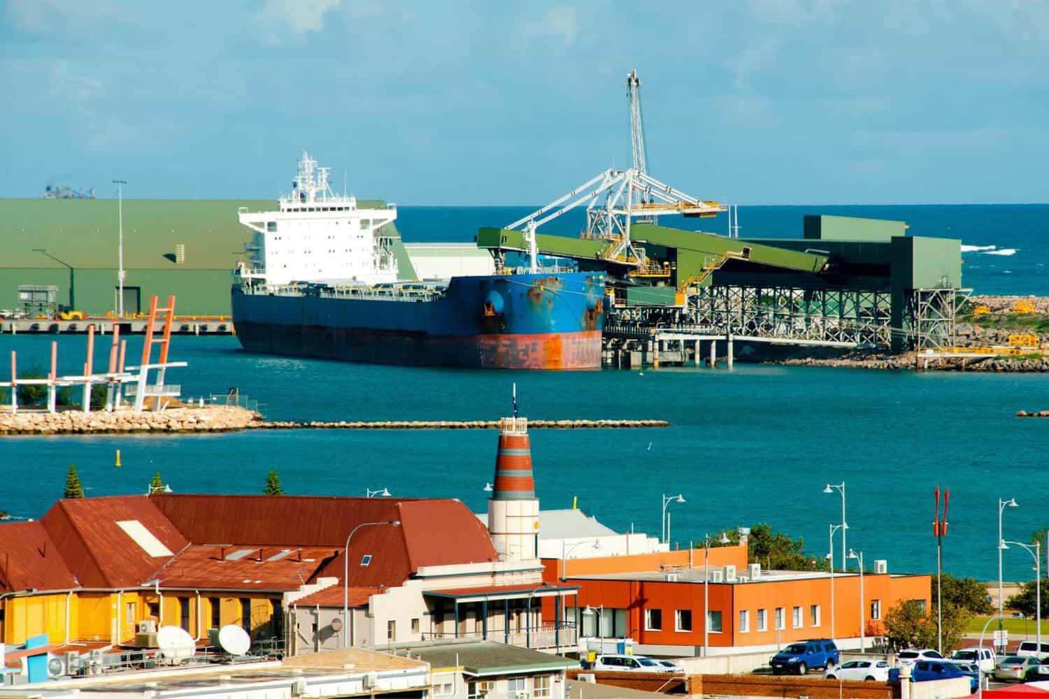 The bustling Geraldton Port with a large cargo ship docked at the iron ore loading terminal, set against a backdrop of vibrant turquoise ocean and a clear sky. 