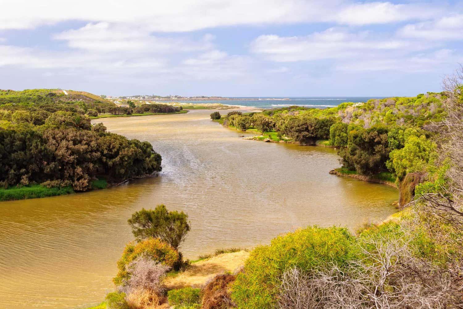 A winding river meanders through dense greenery, leading to the blue ocean in the distance, with a glimpse of the coastal city of Geraldton on the horizon.