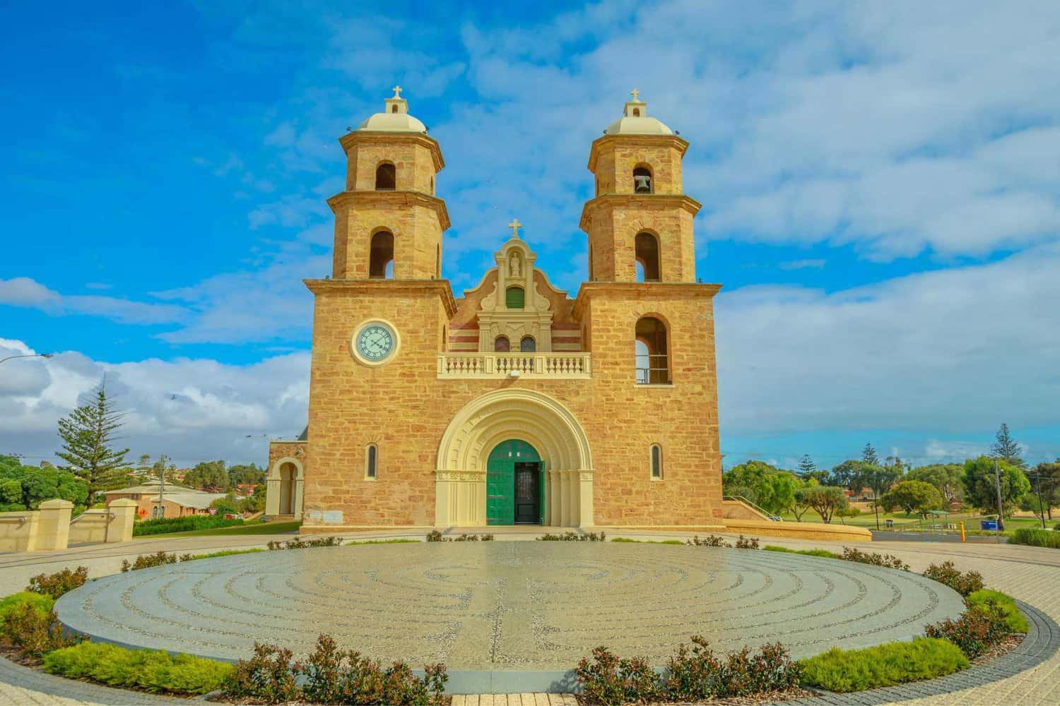 St. Francis Xavier Cathedral in Geraldton, Western Australia, showcasing its distinctive sandy-hued twin towers and central clock under a blue sky with fluffy clouds, a circular patterned forecourt in the foreground.